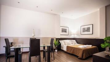 Habitación de hotel con cama, mesita de noche, lámpara y un área de comedor con sillas y una mesa.