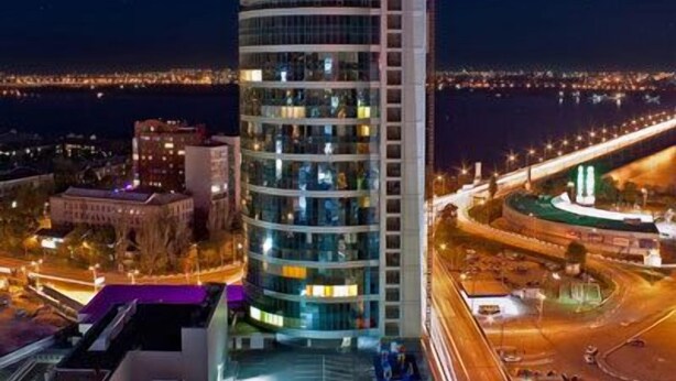 A modern hotel with a cylindrical design, located near a waterfront with a bridge in the background. - Most City Premium apartments (Dnipro)