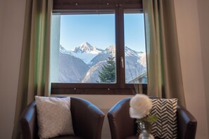A room with a large window overlooking a mountain range. There are two chairs with cushions and a vase with a flower on a table in the foreground. - Hotel Alpenhof (Unterbaech)