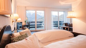 A hotel room with a bed, two bedside lamps, a TV, a desk with a chair, and a view of mountains through large windows. - Hotel Alpenhof (Unterbaech)