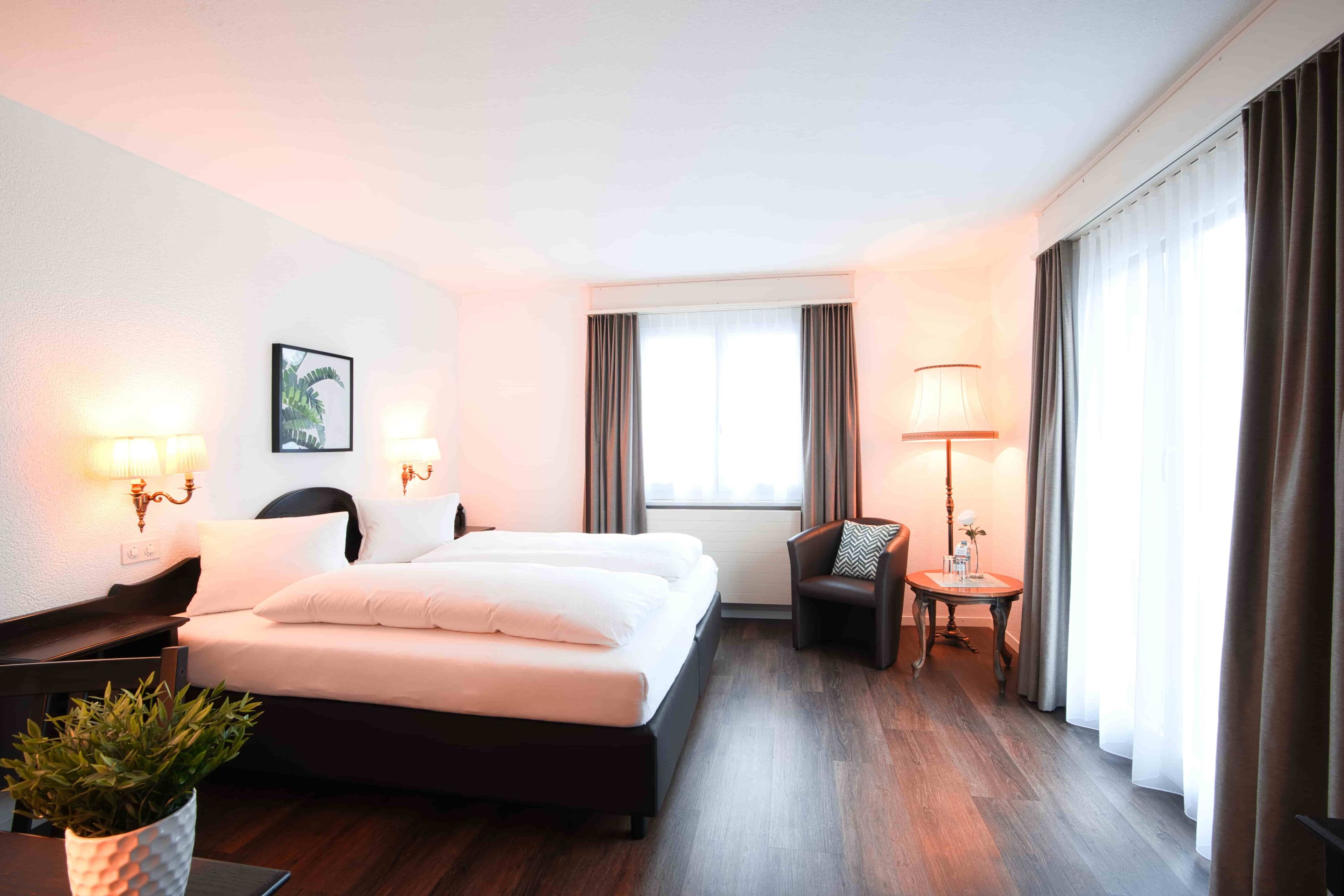Una habitación de hotel con una cama grande, dos lámparas de noche, una mesita con lámpara, una silla y una planta en maceta.