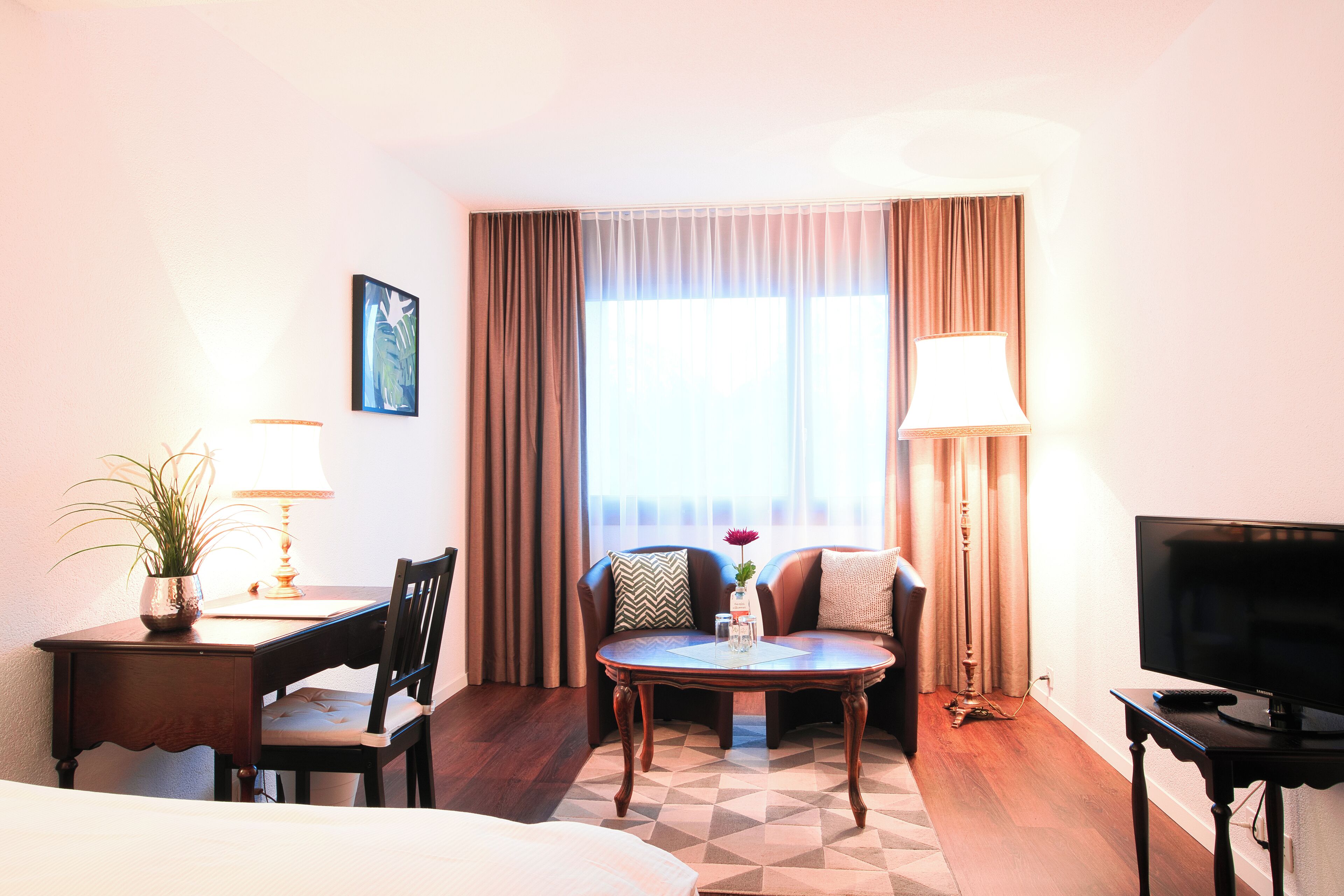 A hotel room with a bed, a desk with a lamp, a chair, a table with a vase, a television, and a window with curtains.
