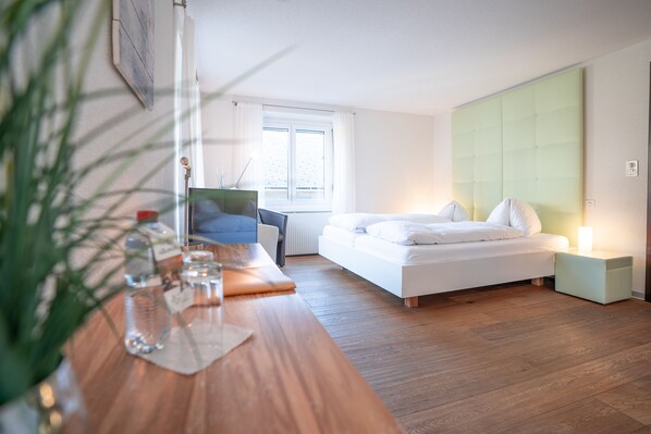 A modern bedroom with a wooden desk, a bed with white bedding, a green headboard, and a window with white curtains. - Hotel Alpenhof (Unterbaech)
