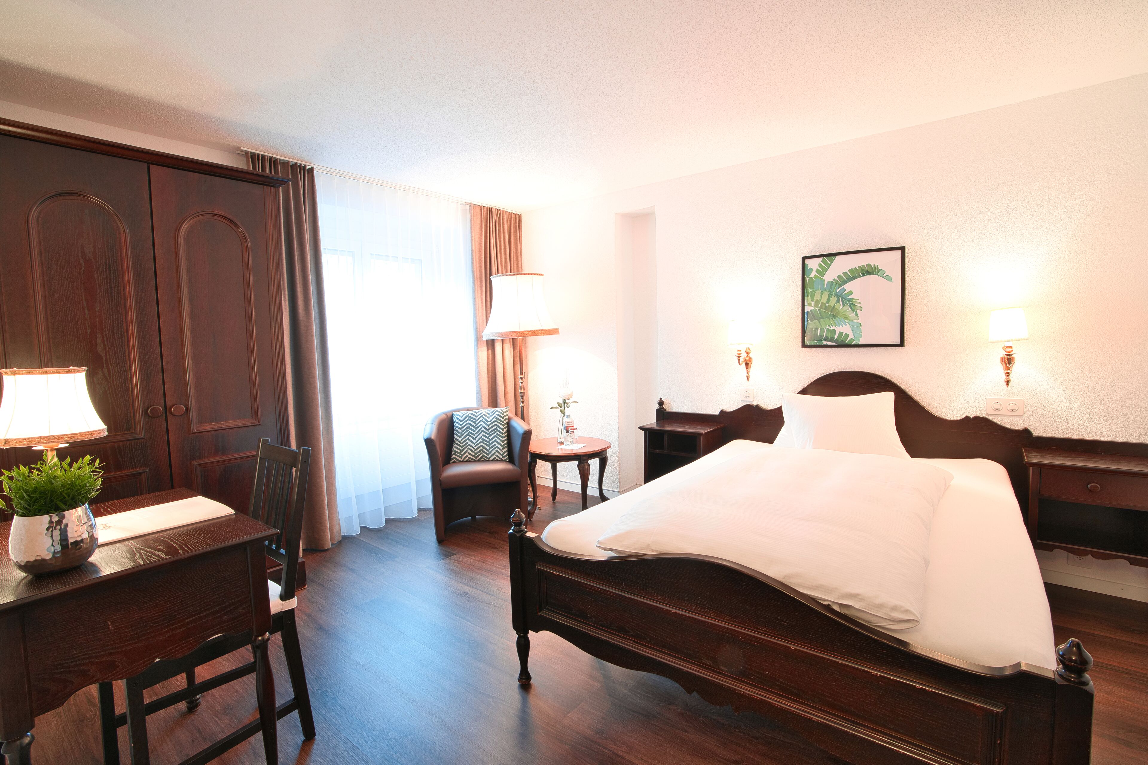 A hotel room with a wooden bed, a desk with a lamp, a chair, and a small table with a plant.