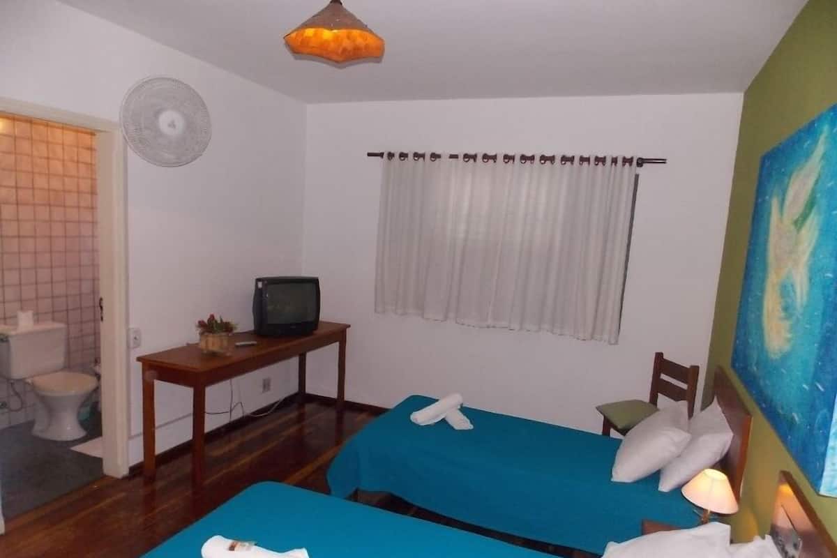Una habitación de hotel con dos camas, una mesita con televisor, una ventana con cortinas y un cuadro en la pared.