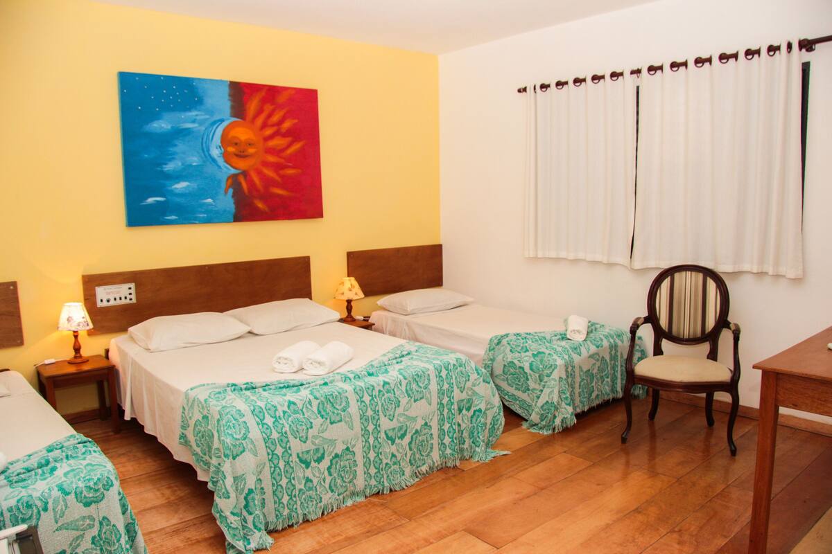 Habitación de hotel con dos camas, una silla de madera, un escritorio, un cuadro en la pared y una ventana con cortinas.