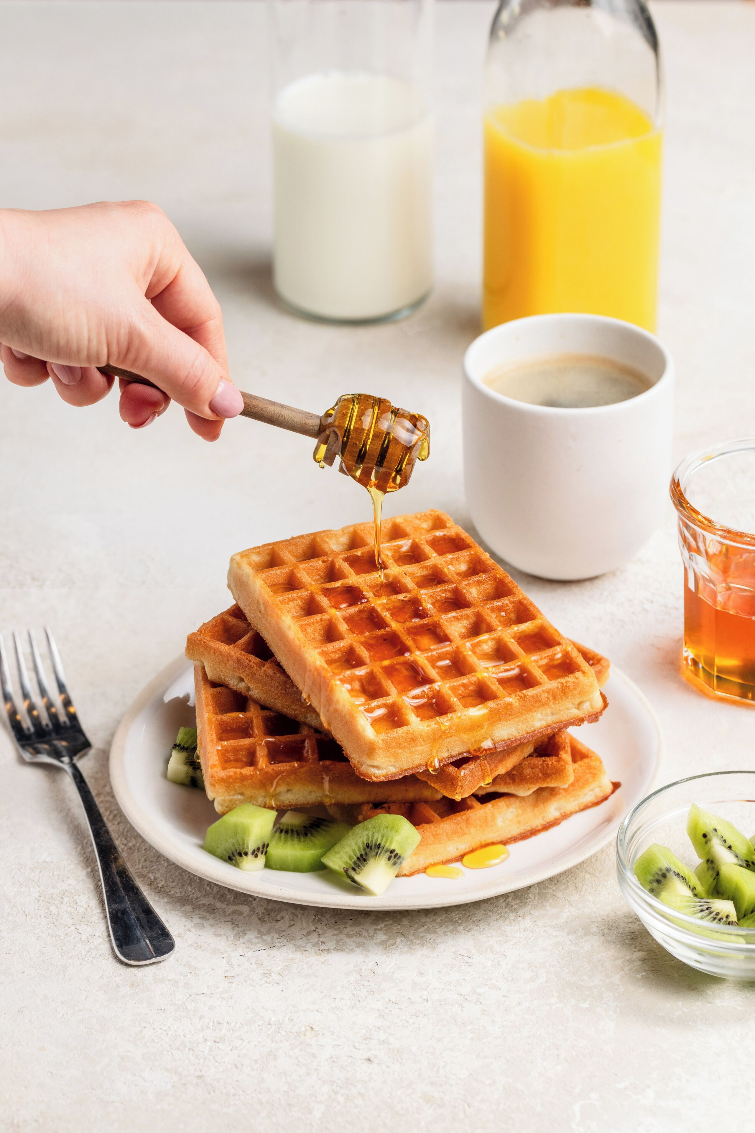 Um prato de waffles com calda e fatias de kiwi, um copo de suco de laranja e uma garrafa de leite ao fundo.
