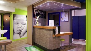 A reception area with a reception desk, a wall with a large portrait, a green mat on the floor, and a blue wall with a poster. - Hotel Ibis Budget Metz Sud Augny (Augny)