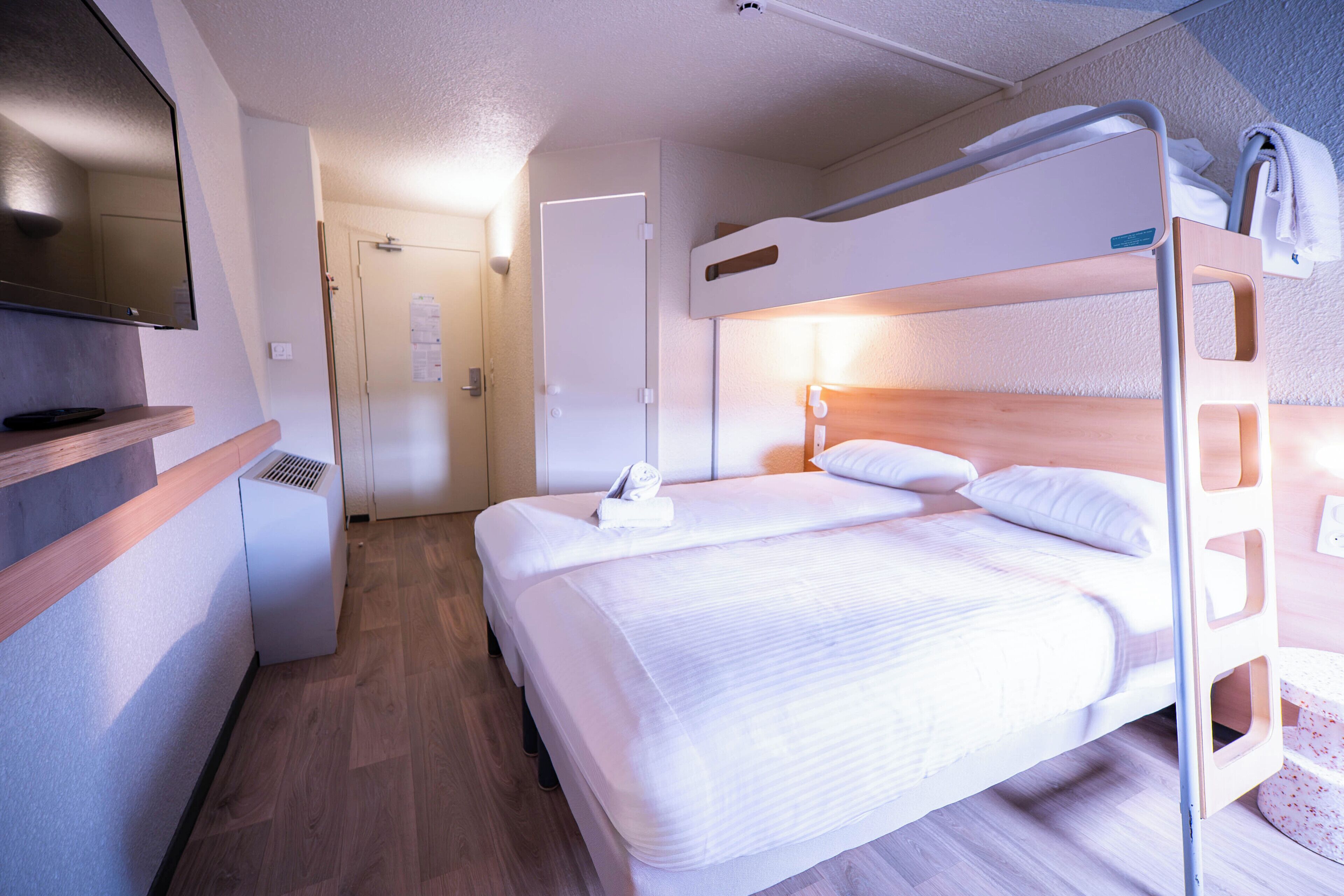 A hotel room with a bunk bed, a flat-screen TV, a desk, and a wooden floor.