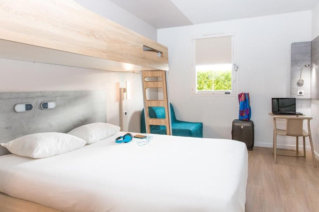 Ein Hotelzimmer mit Etagenbett, Schreibtisch mit Laptop, Stuhl, Koffer und Fenster mit Blick ins Grüne.