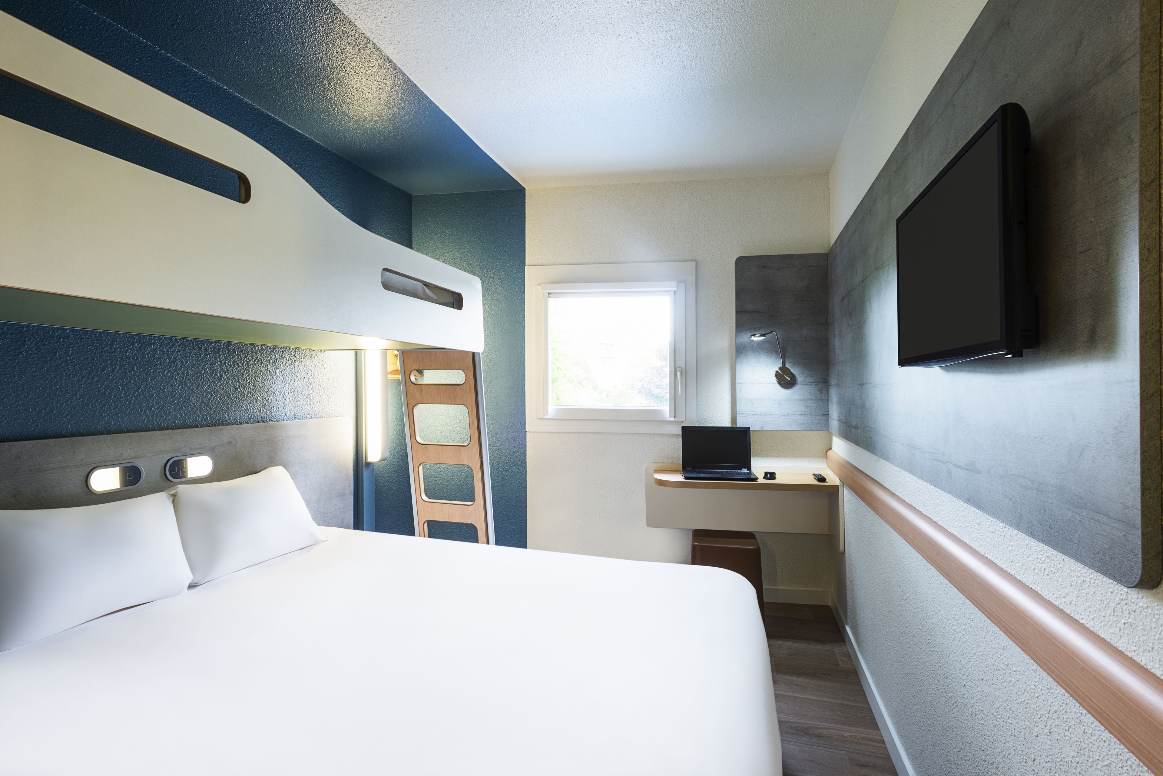 A modern hotel room with a large bed, a desk with a laptop, a flat-screen TV mounted on the wall, and a window providing natural light.