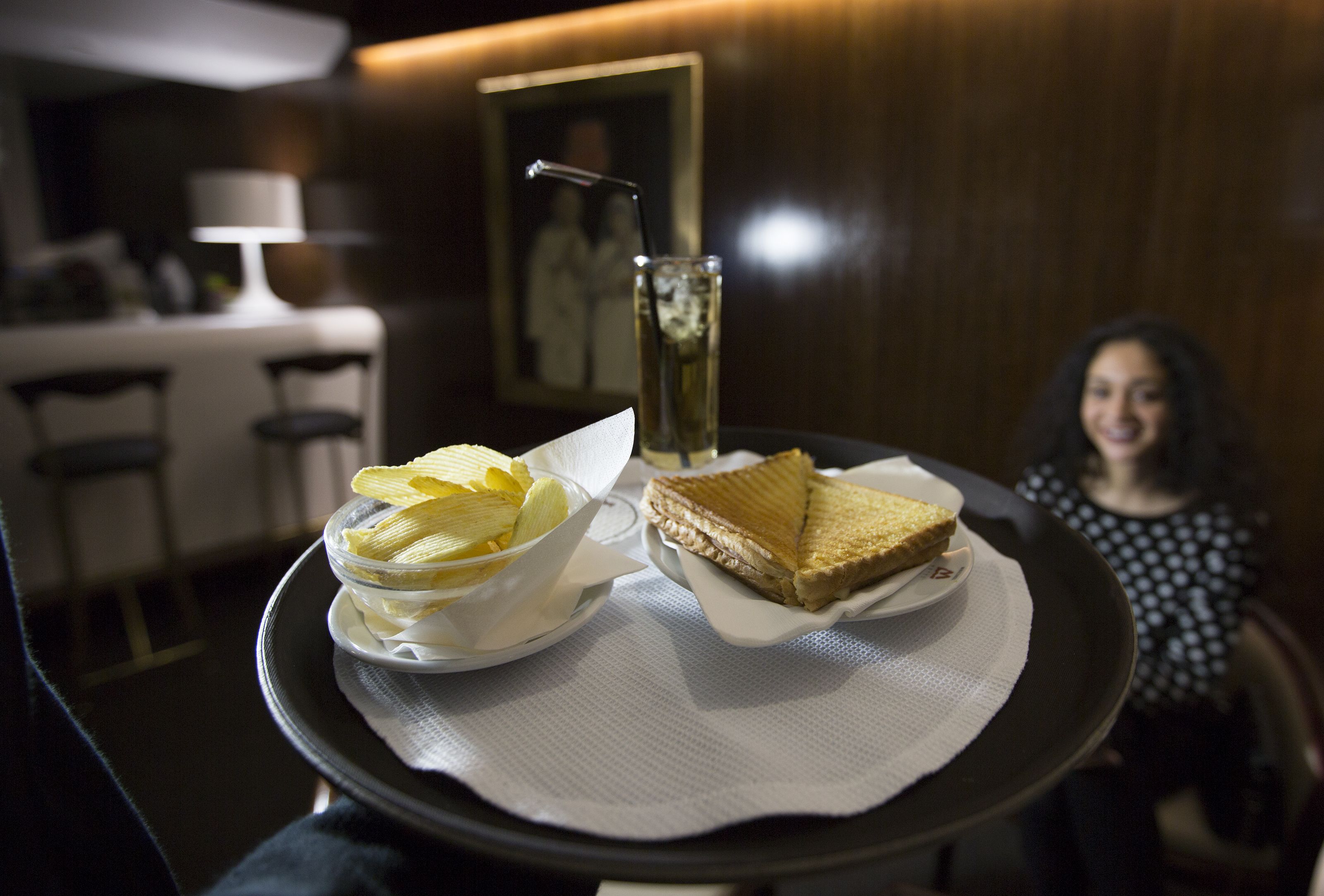 Una bandeja con un sándwich y papas fritas sobre una mesa en una habitación con poca luz, con un comedor al fondo.