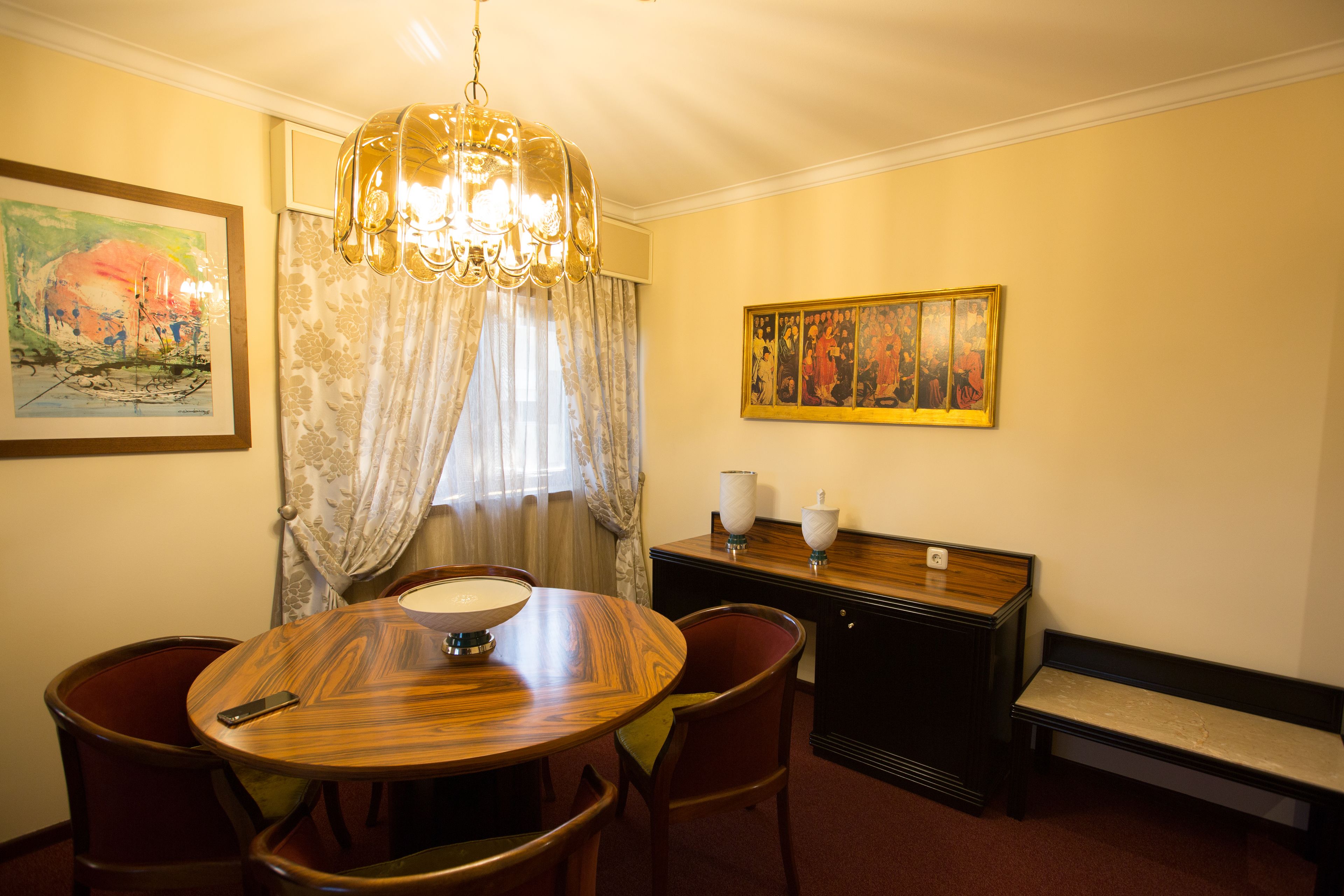 A dining area with a round wooden table, chairs, a chandelier, a painting, and a cabinet.