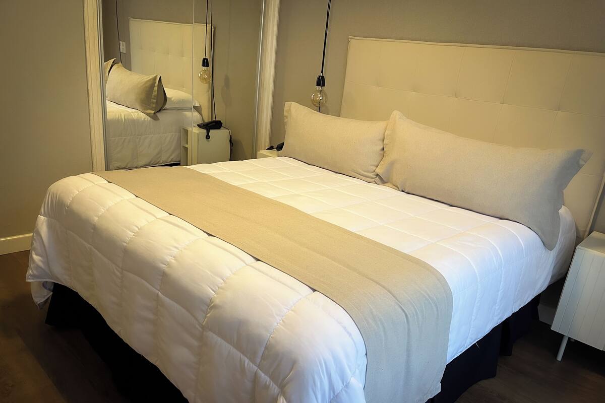A neatly made bed with a white duvet and a beige throw blanket, a bedside table with a lamp, and a mirror on the wall.