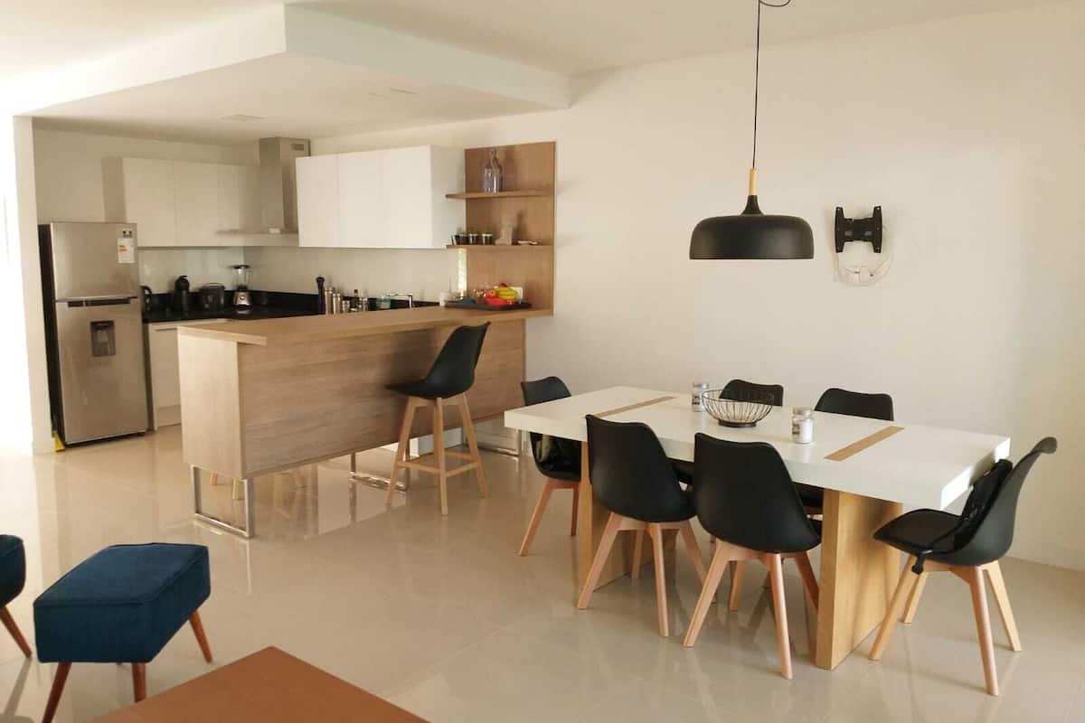 A modern kitchen with a central island, a dining area with six chairs, and a black pendant light.