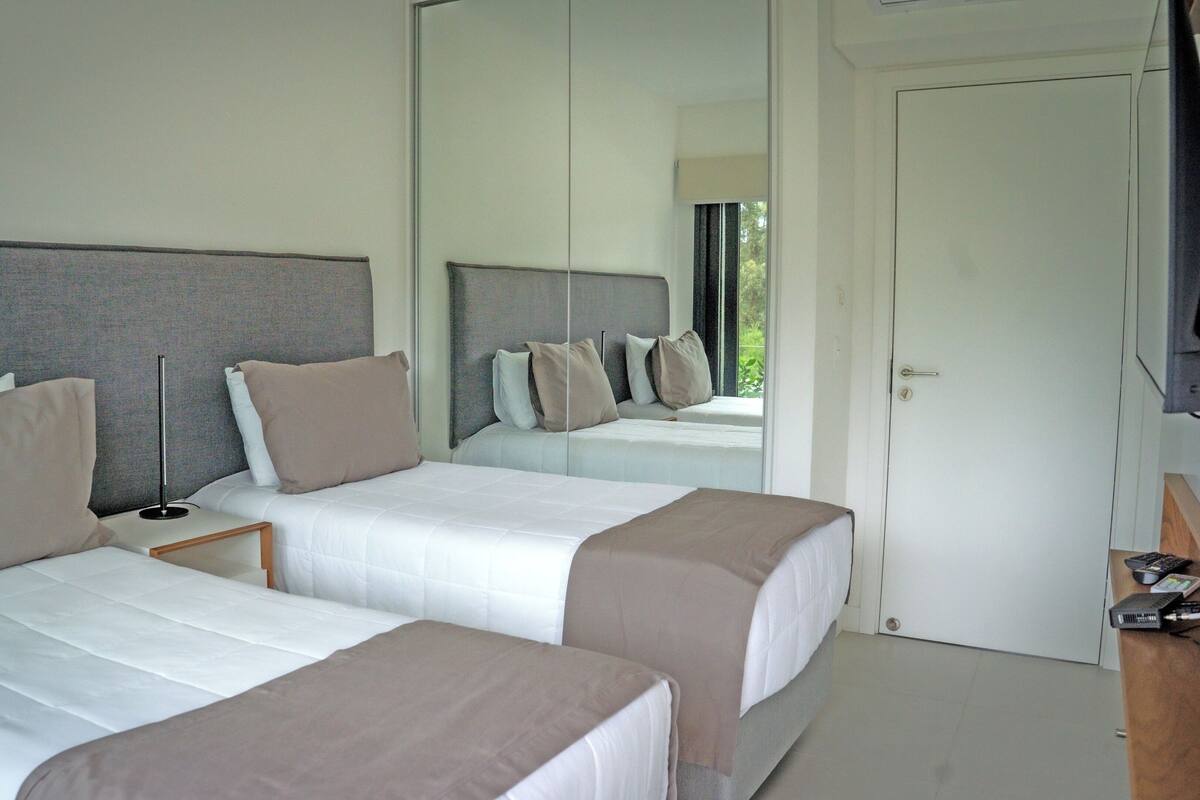 A hotel room with two beds, a bedside table with a lamp, a mirror, and a door.