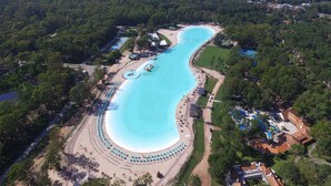 An aerial view of a resort with a large, curved swimming pool, surrounded by beach areas with umbrellas and lounge chairs.