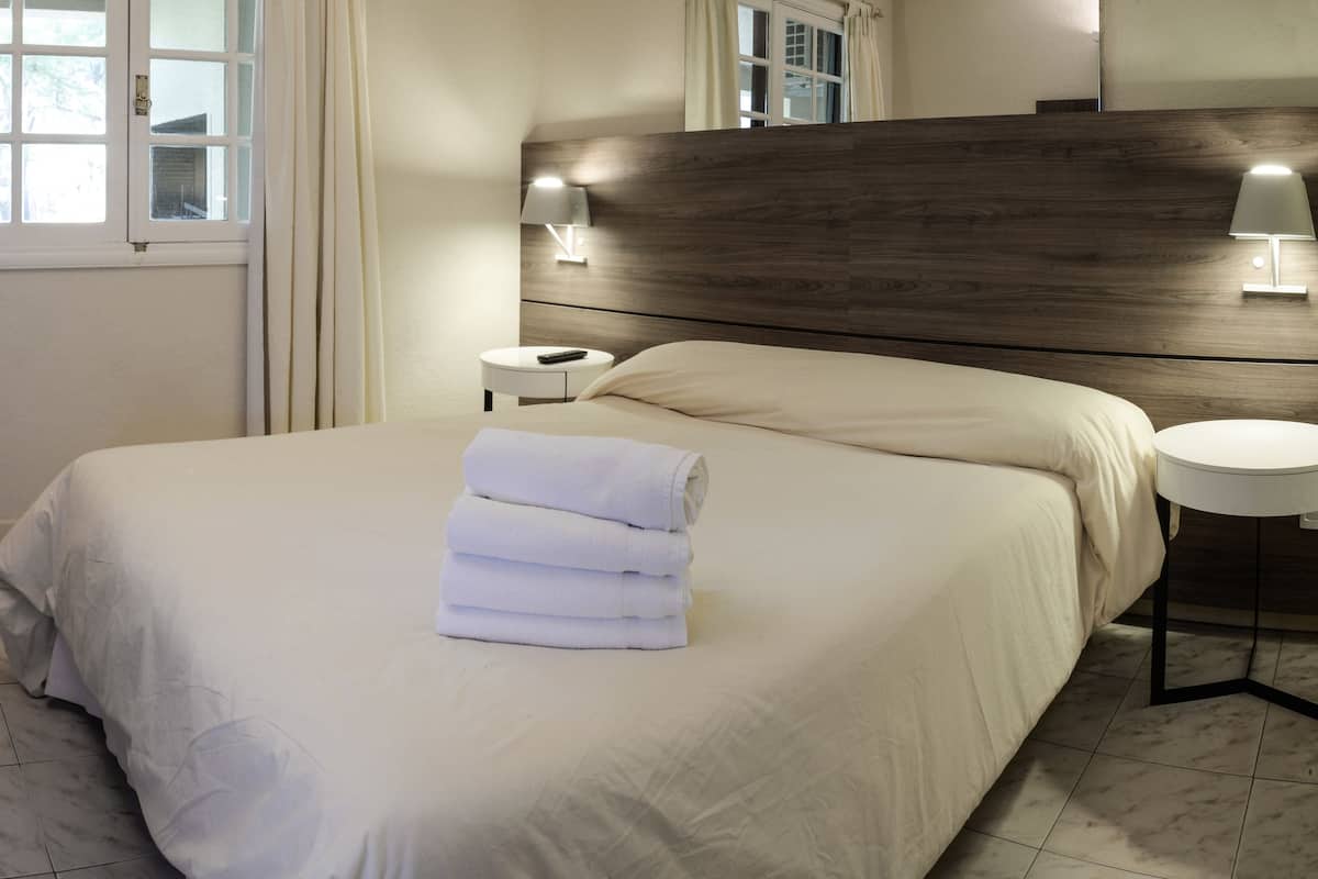 A hotel room with a neatly made bed, a bedside table with a lamp, and a window with curtains.