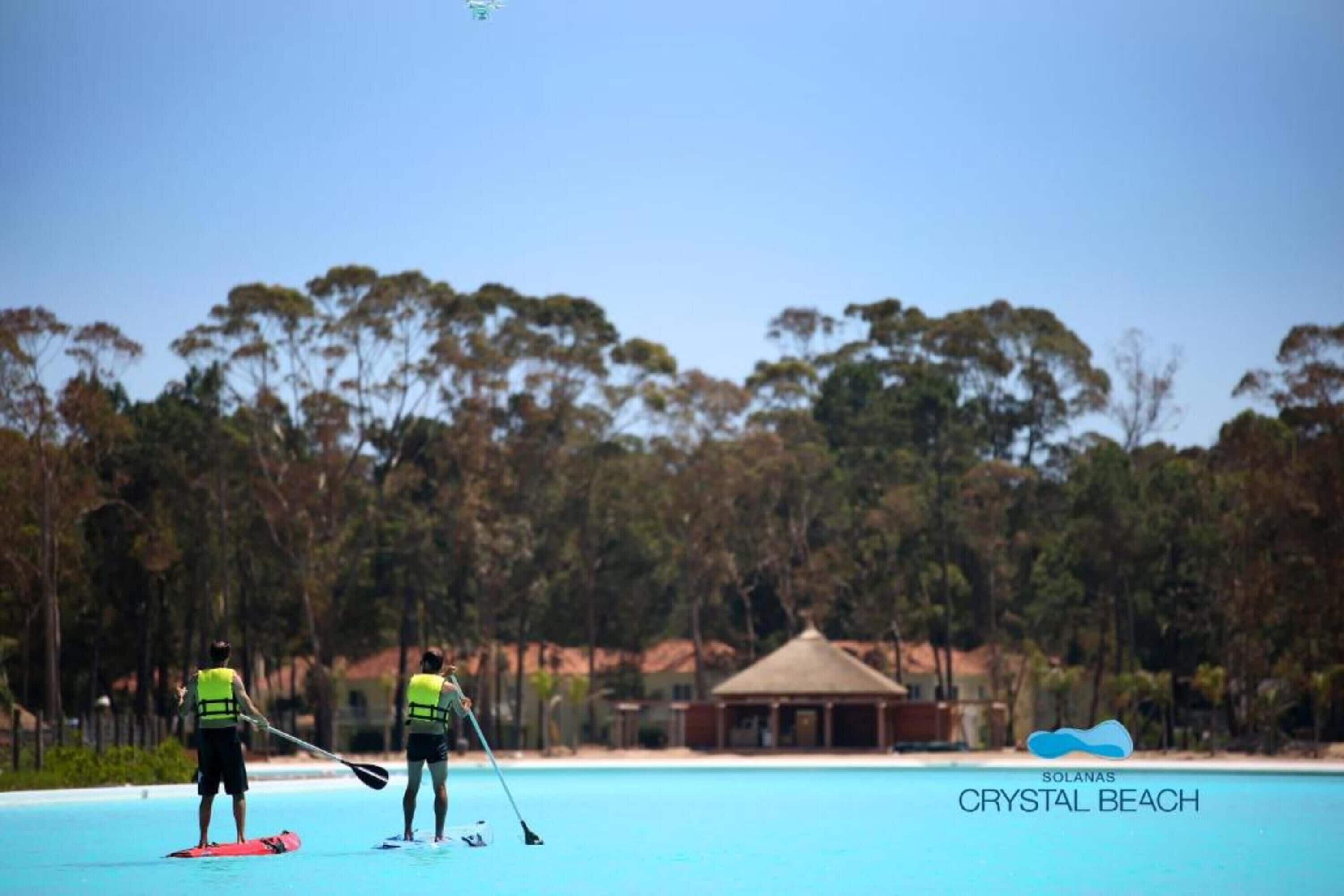 Duas pessoas com coletes salva-vidas em uma prancha de remo em uma piscina azul cristalina, com uma casa de praia e árvores ao fundo.