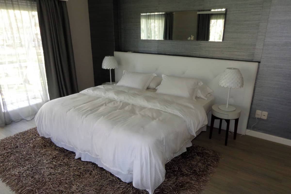 A modern bedroom with a large bed, a bedside lamp, a mirror above the bed, and a view of the outdoors through a window.