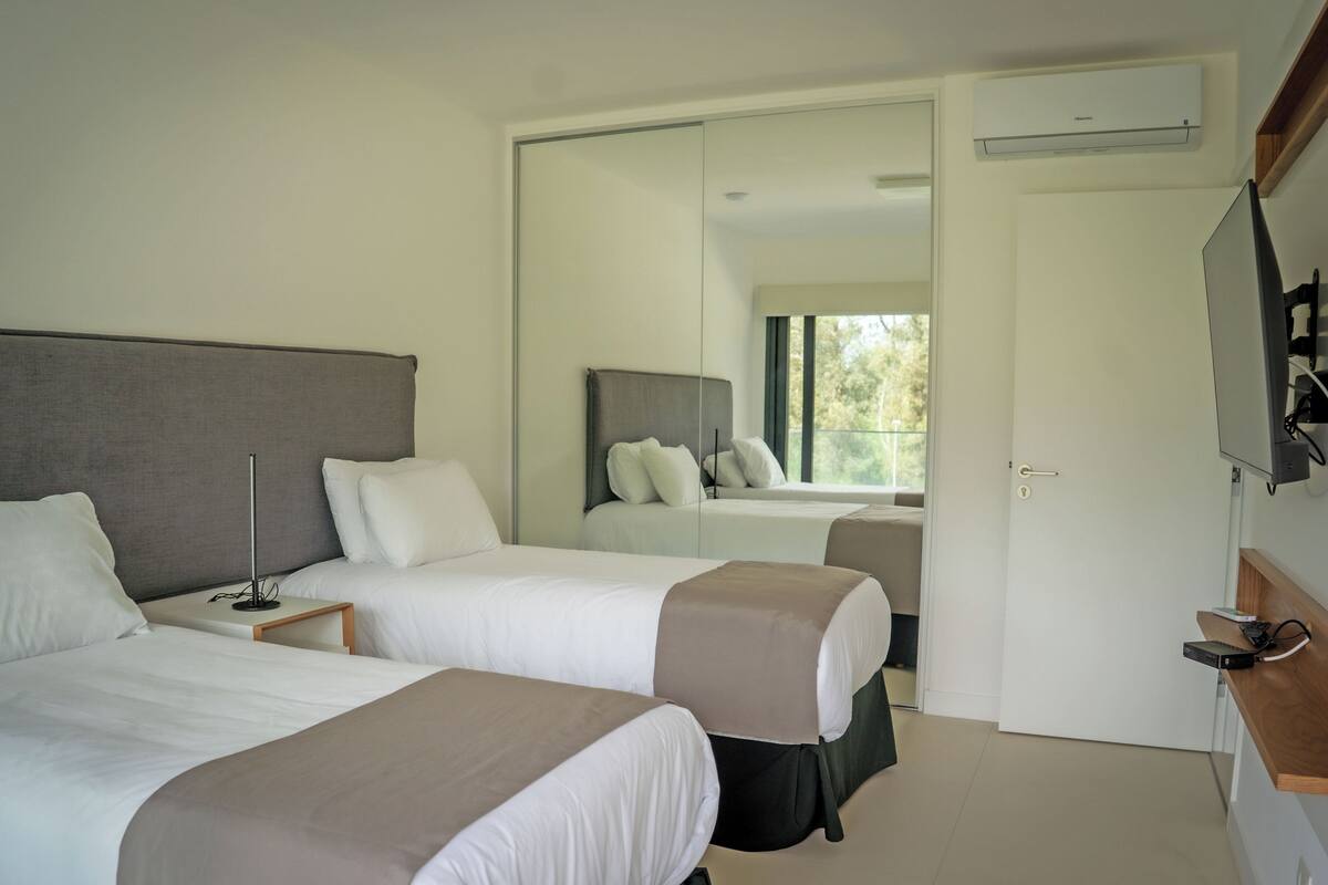 A hotel room with two beds, a bedside table with a lamp, a large mirror, and an air conditioning unit.