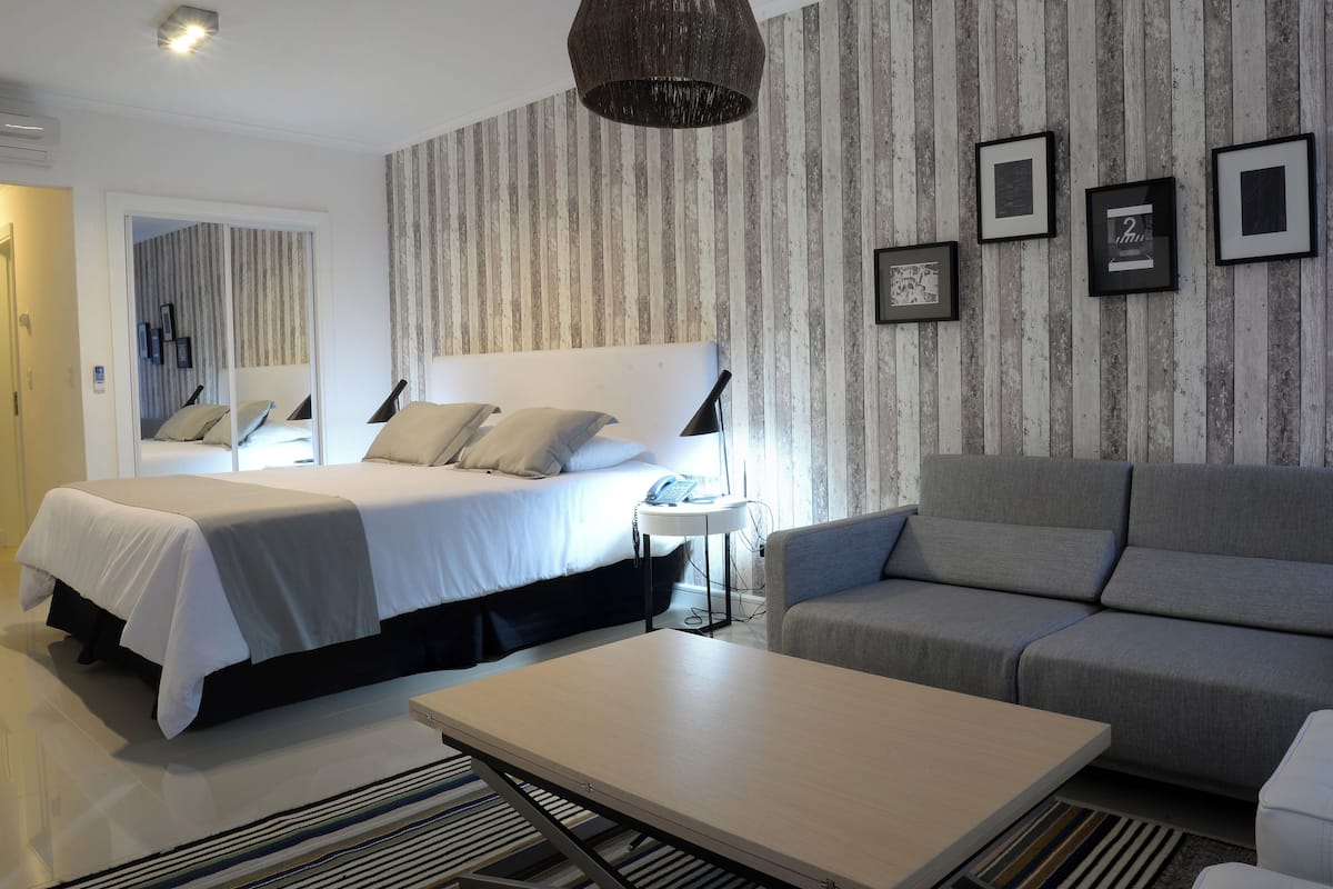 A modern hotel room with a large bed, a grey sofa, a small table, and a wall with striped wallpaper and framed pictures.