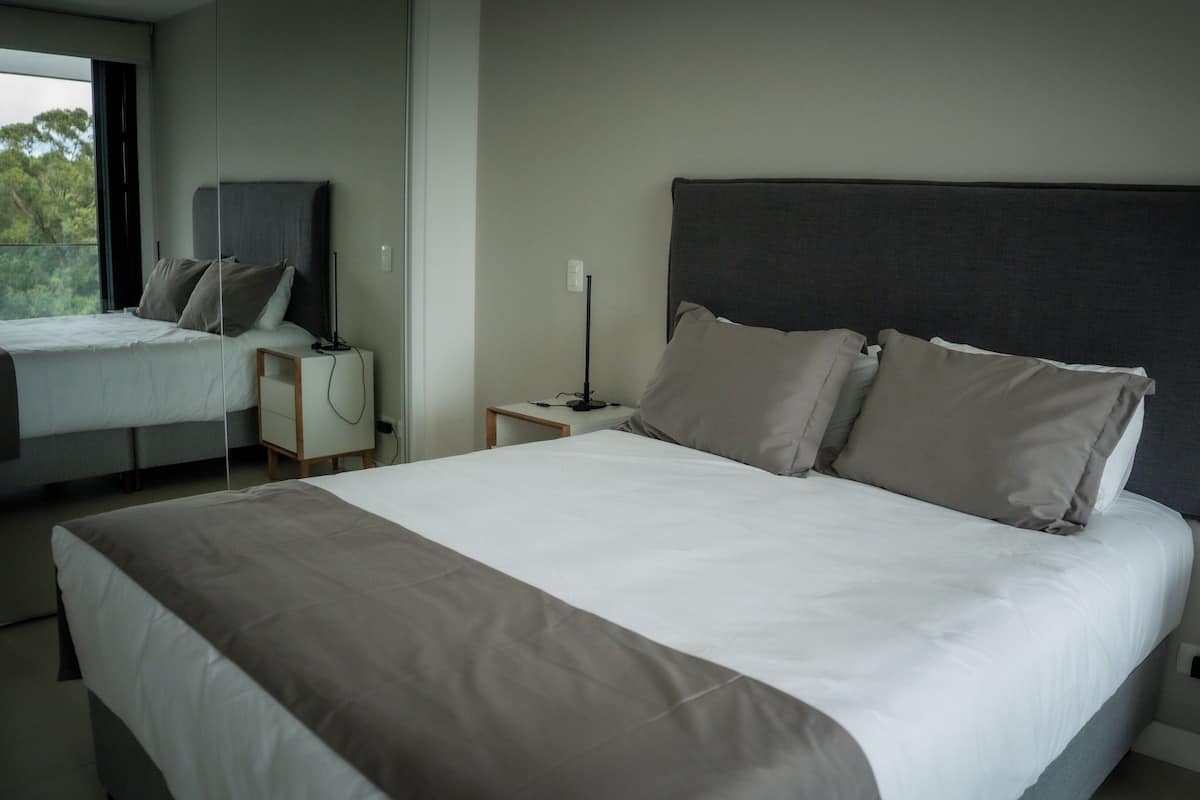 A modern hotel room with a large bed, a bedside table, a nightstand, and a window with a view of trees.