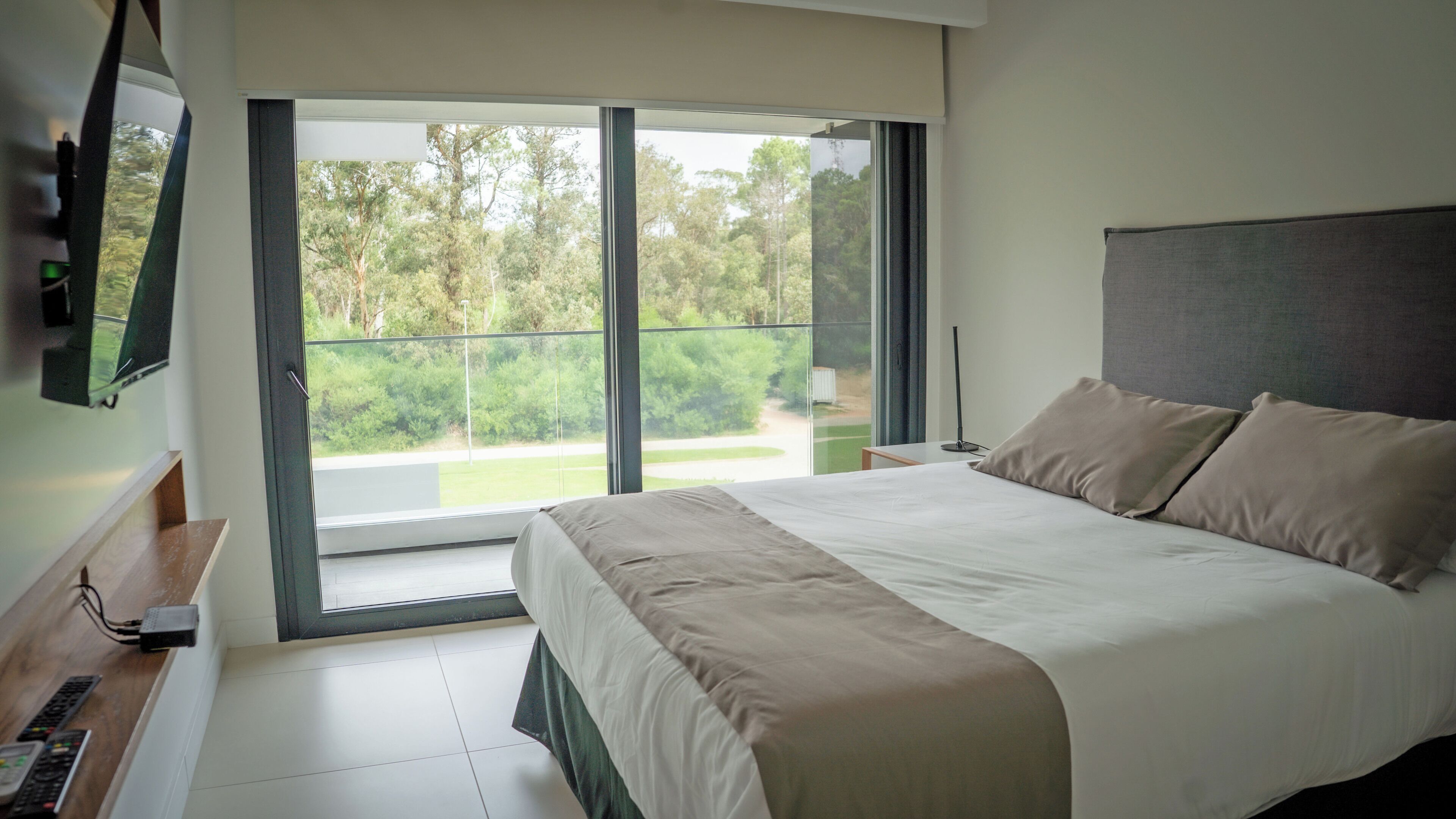 Un dormitorio moderno con una cama grande, un televisor de pantalla plana en la pared y un ventanal corredizo que da a un balcón con vistas a zonas verdes.