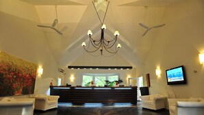 A hotel lobby with a reception desk, two white sofas, a chandelier, ceiling fans, and a television on the wall.