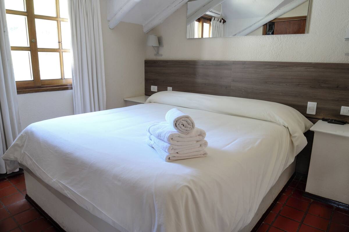 A neatly made bed with a rolled towel on top, a bedside table, a nightstand, a lamp, and a window with curtains.