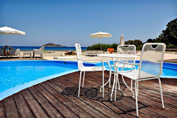 A poolside area with white metal chairs and tables, umbrellas, and a view of the sea.