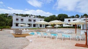 A pool area with lounge chairs and umbrellas, a white building with multiple balconies, and a clear sky. - Tony's Beach (Leros)