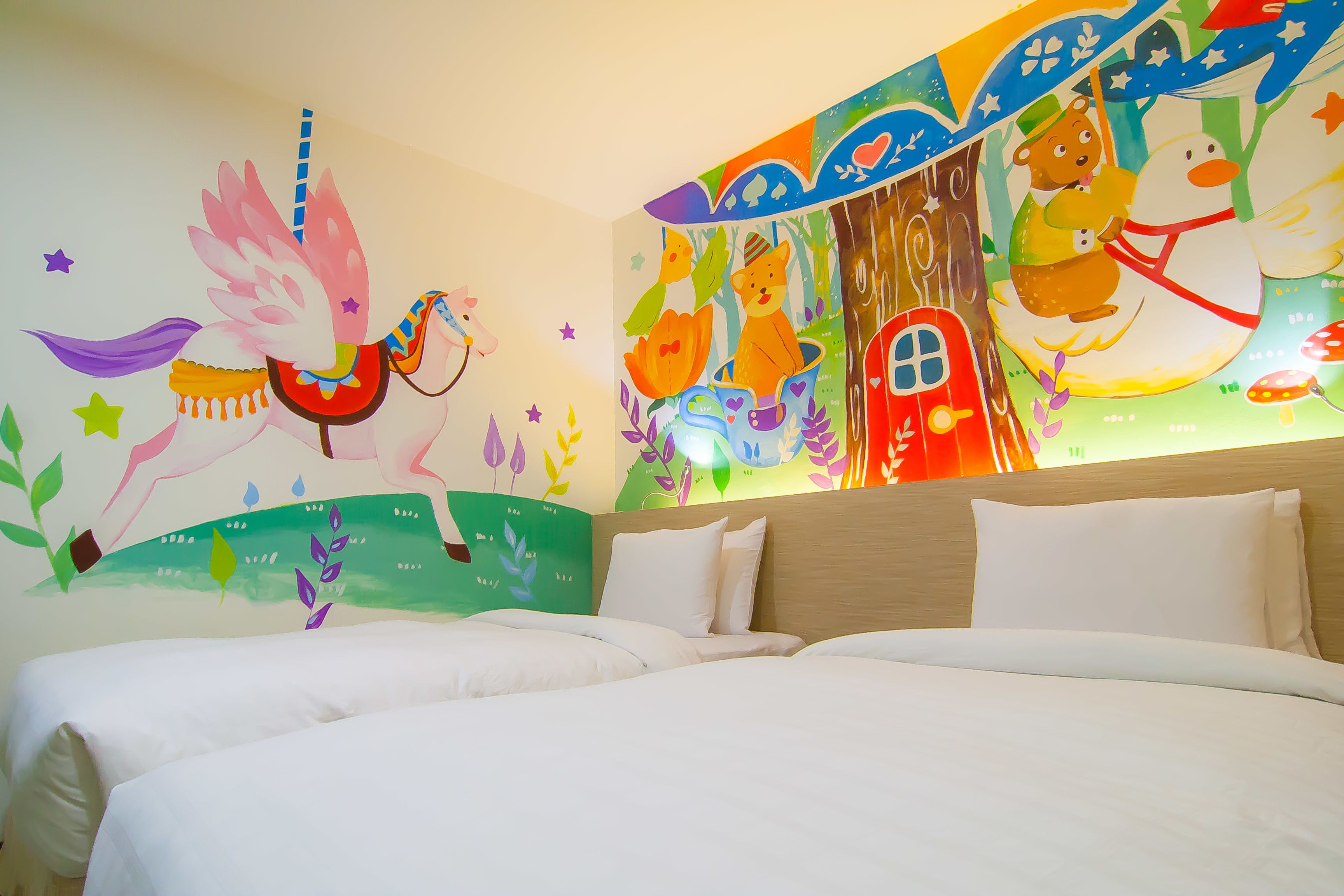 Habitación de hotel con una cama grande, dos almohadas y un mural de colores en la pared que representa animales y una casa.