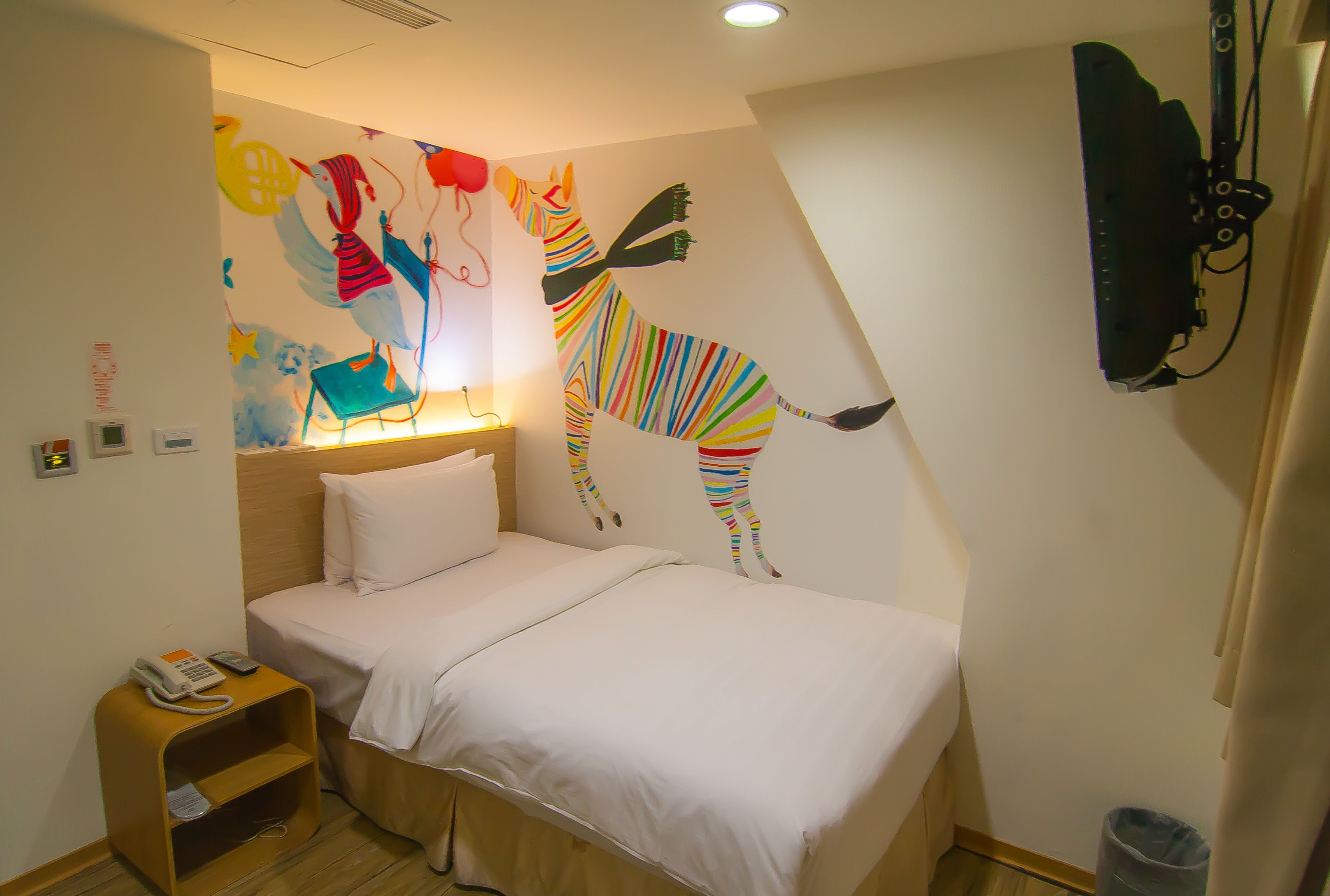 Een hotelkamer met een bed, een nachtkastje, een wandtelevisie en een kleurrijke muurschildering.