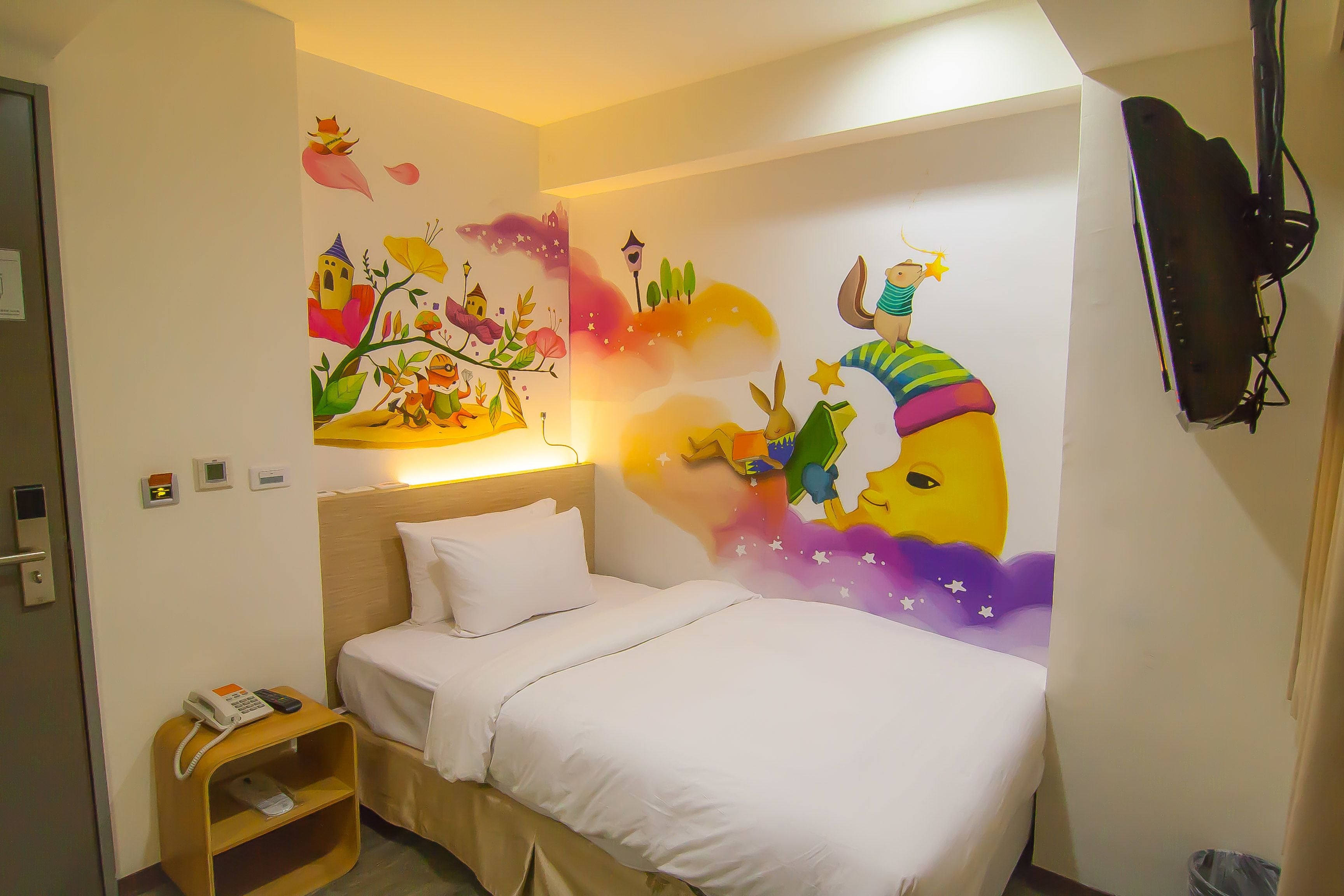 A hotel room with a bed, a bedside table with a phone, a wall-mounted TV, and a colorful mural on the wall.
