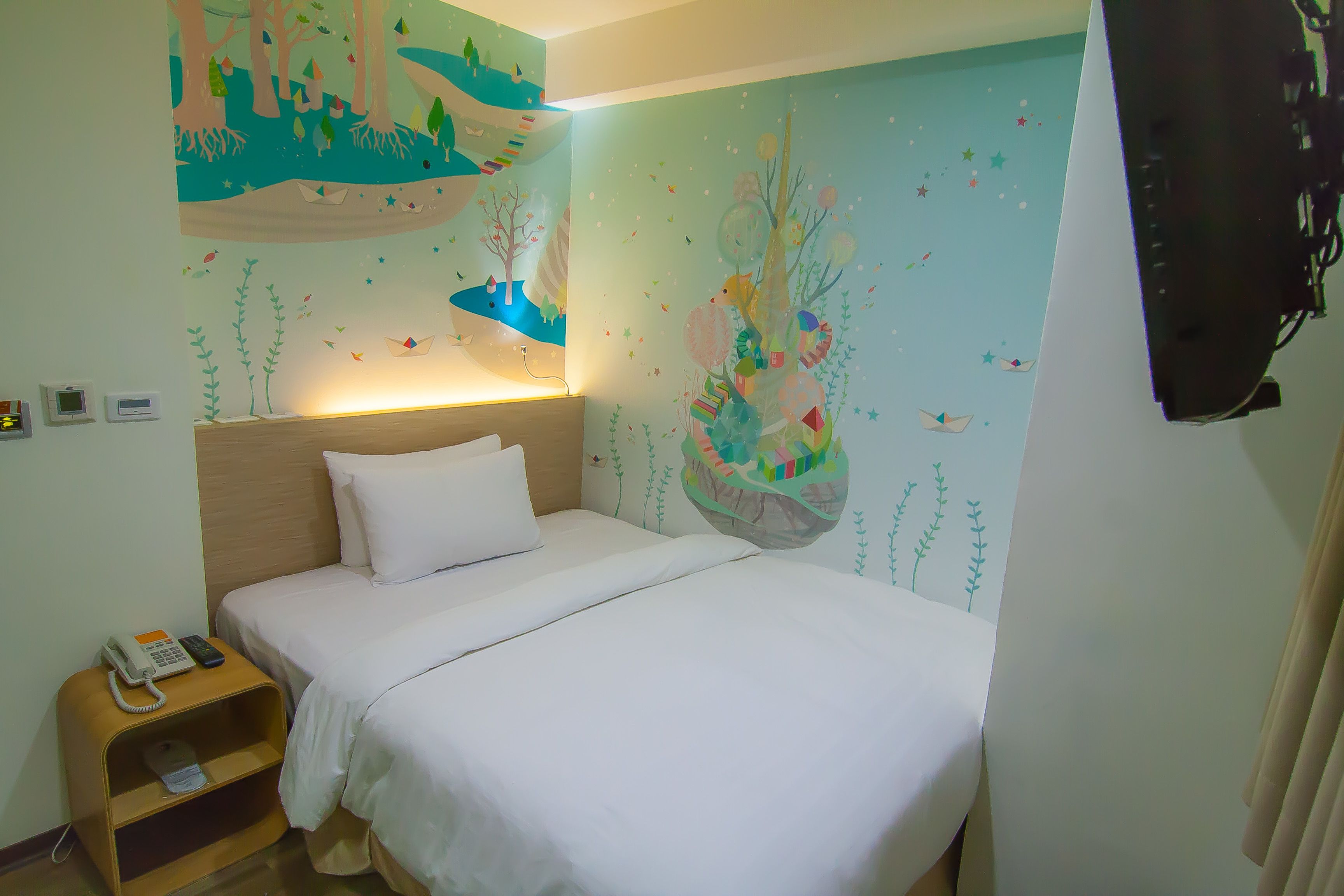 A hotel room with a bed, a bedside table with a phone, a wall mural, and a hanging basket.