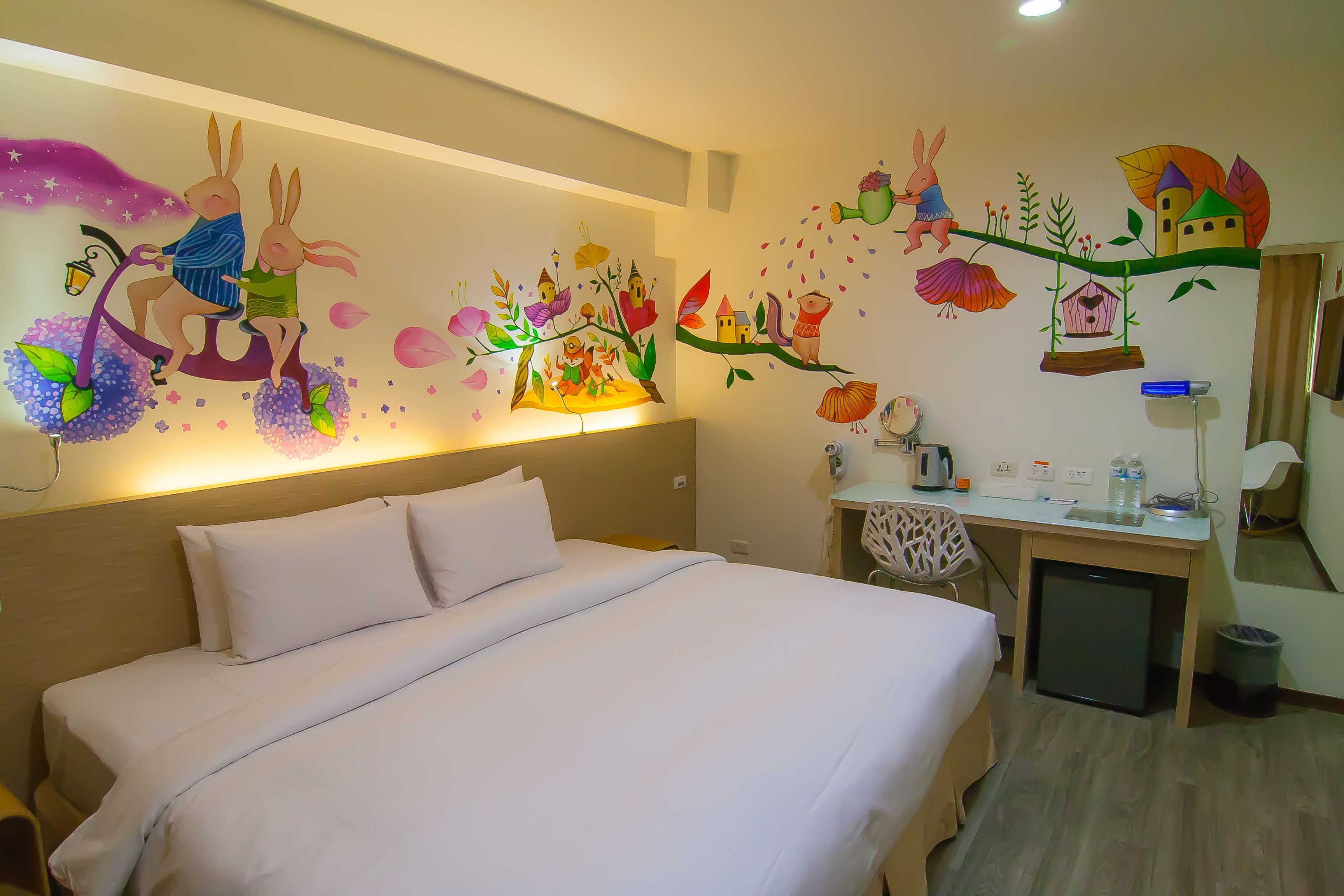 Una habitación de hotel con una cama, un escritorio con silla, un refrigerador pequeño y una pared con murales coloridos.