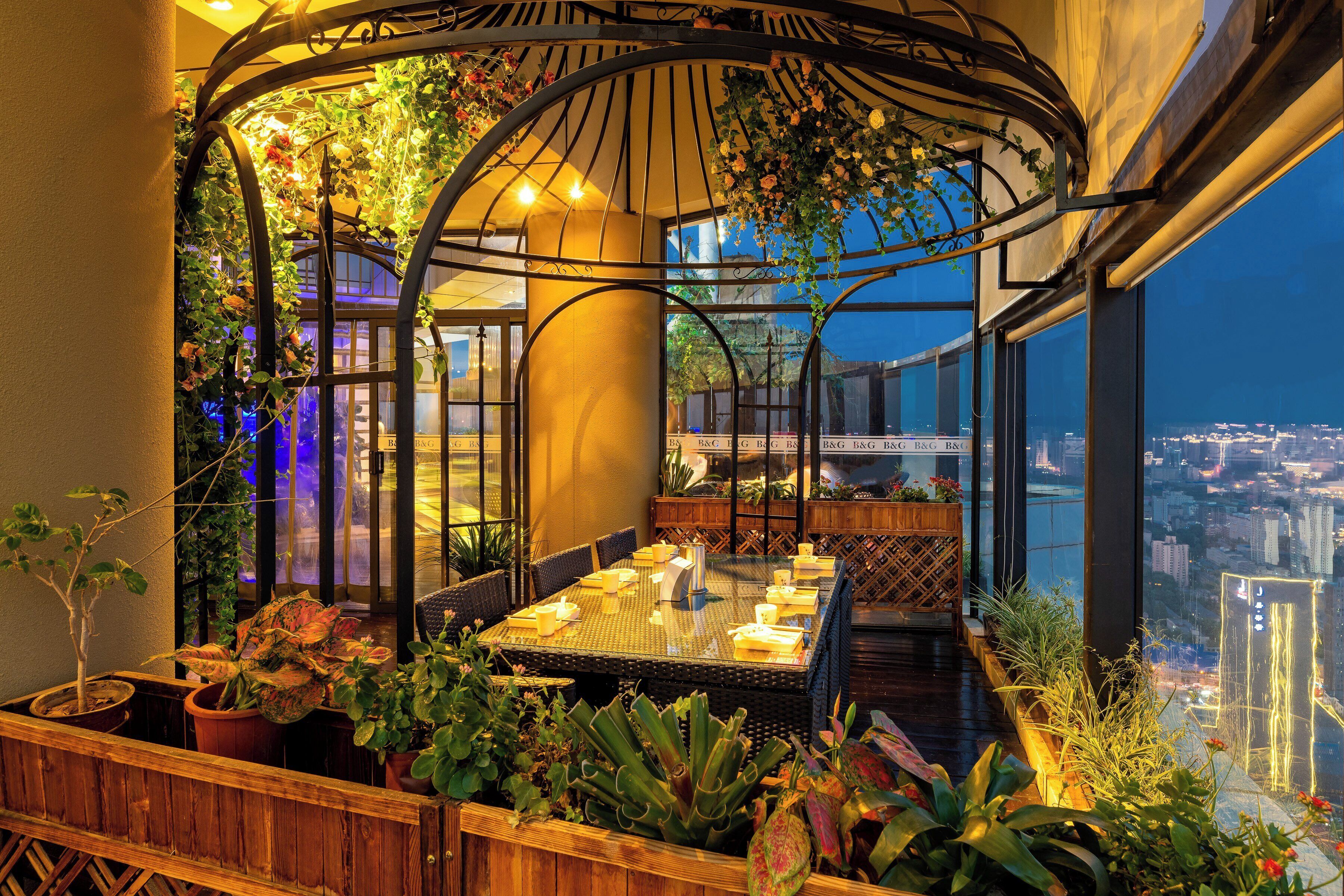 Terraza en la azotea con una mesa de vidrio dispuesta para comer, rodeada de plantas en macetas y una estructura de pérgola de metal.