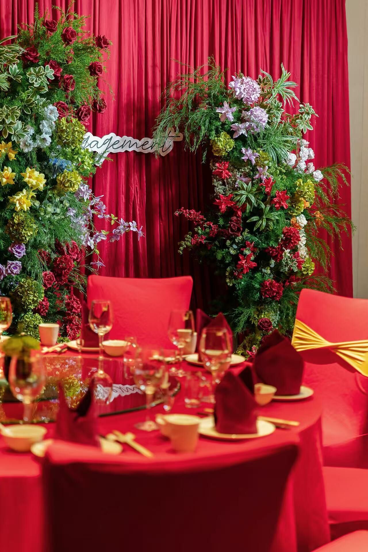 A dining table set for a meal with red chairs, a red tablecloth, and a floral arrangement in the background.