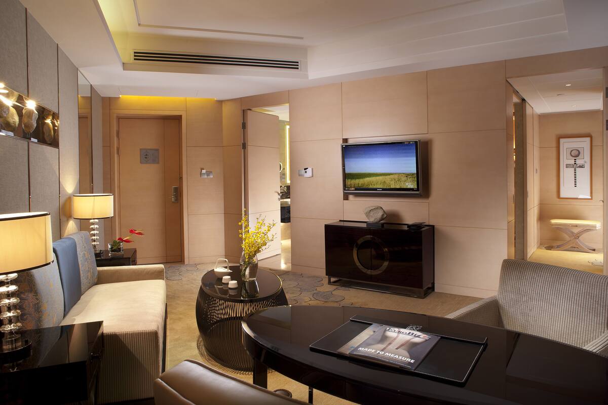 A modern hotel room with a flat-screen TV mounted on the wall, a sofa, a dining table, and a small side table with a vase of flowers.