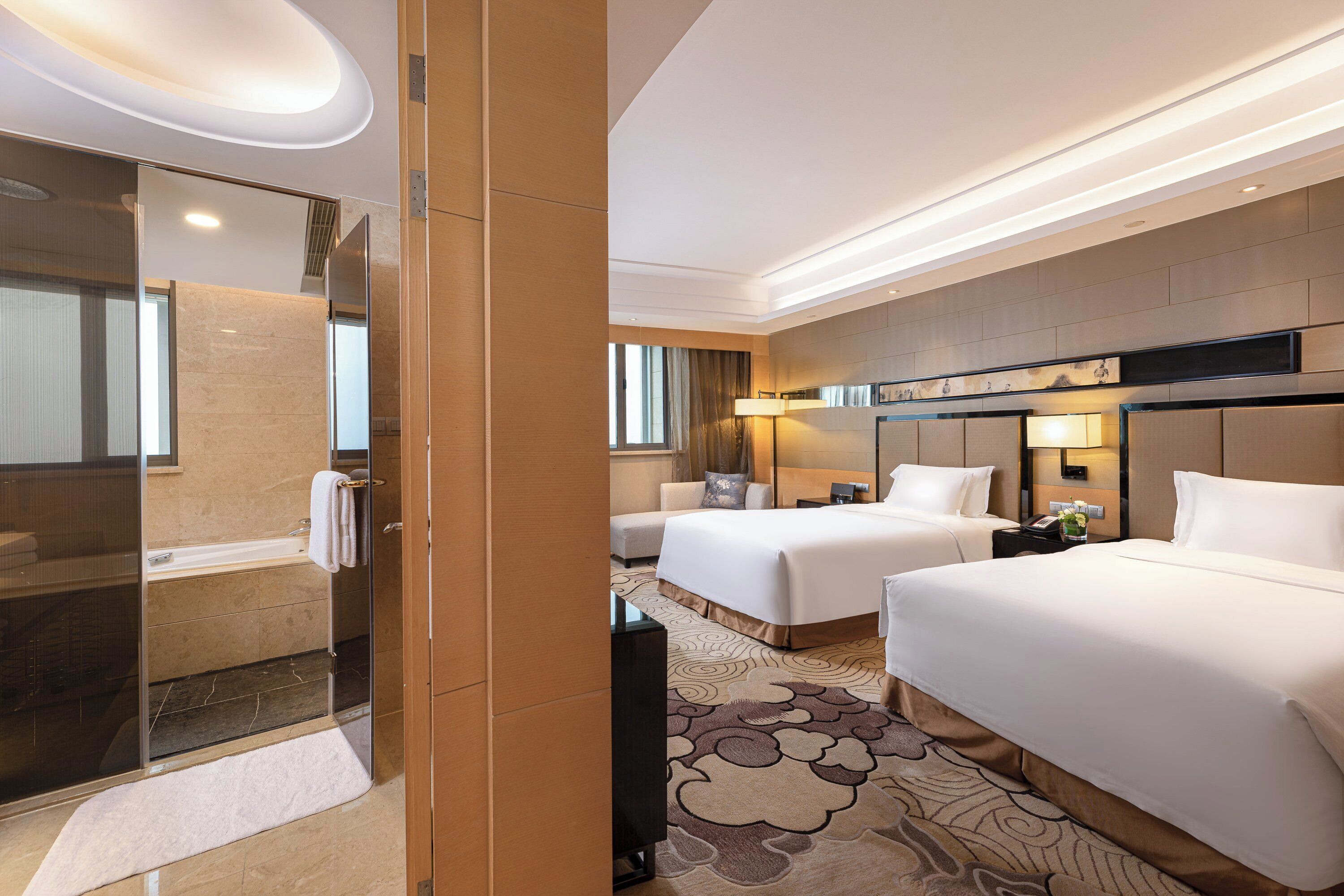Habitación de hotel con cabina de ducha de vidrio, dos camas, una mesita de noche con lámpara y un espejo grande.