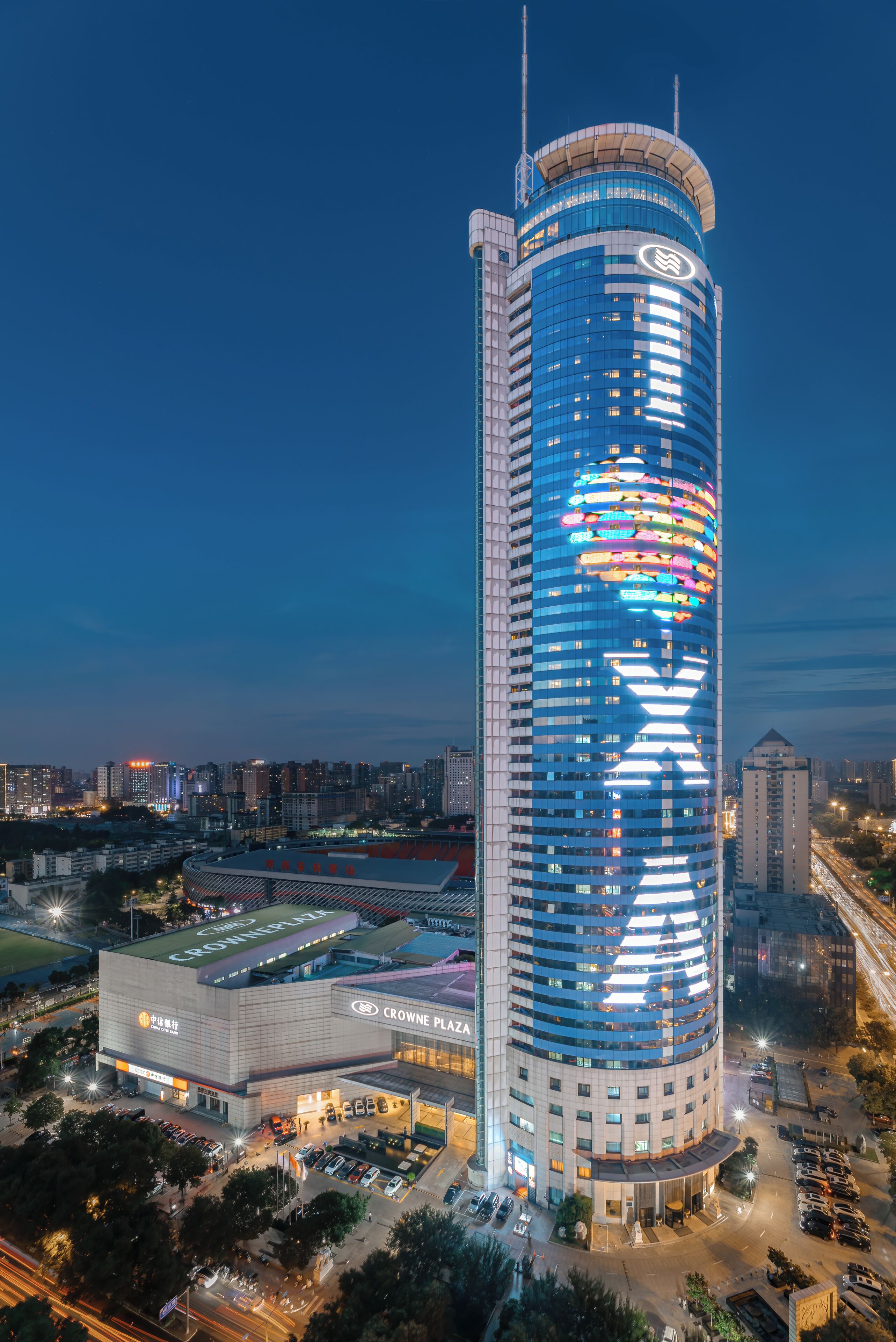 Ein Hochhaus mit einer markanten X-förmigen Lichtinstallation an der Fassade, umgeben von einer Stadtlandschaft in der Dämmerung.
