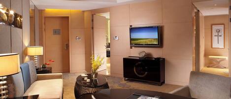 Habitación de hotel con televisor montado en la pared, una mesa de comedor con una revista, un sofá y una mesita auxiliar con un jarrón de flores.