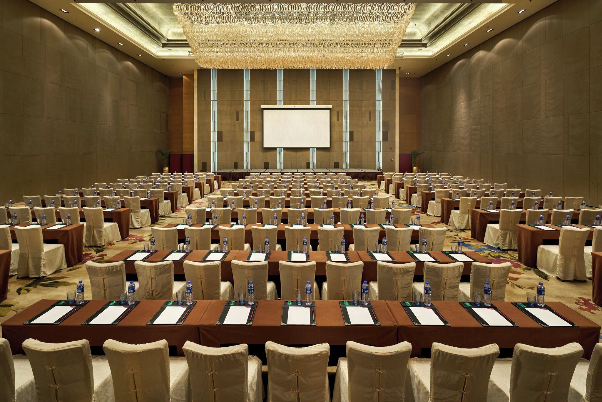 Un salón de conferencias con filas de mesas y sillas, una pantalla grande al frente y una lámpara decorativa en el techo.