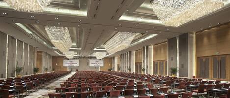 A spacious conference room with rows of tables and chairs, large chandeliers, and a high ceiling.