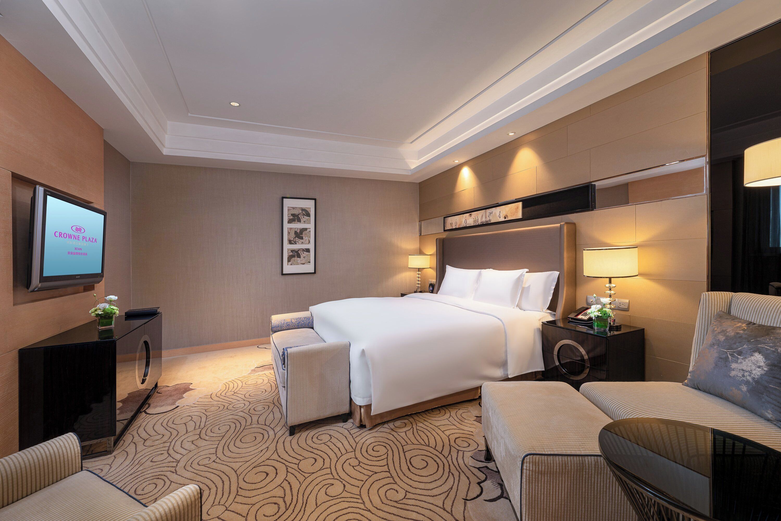 A hotel room with a large bed, a desk with a computer, a chair, a sofa, and a TV on the wall.