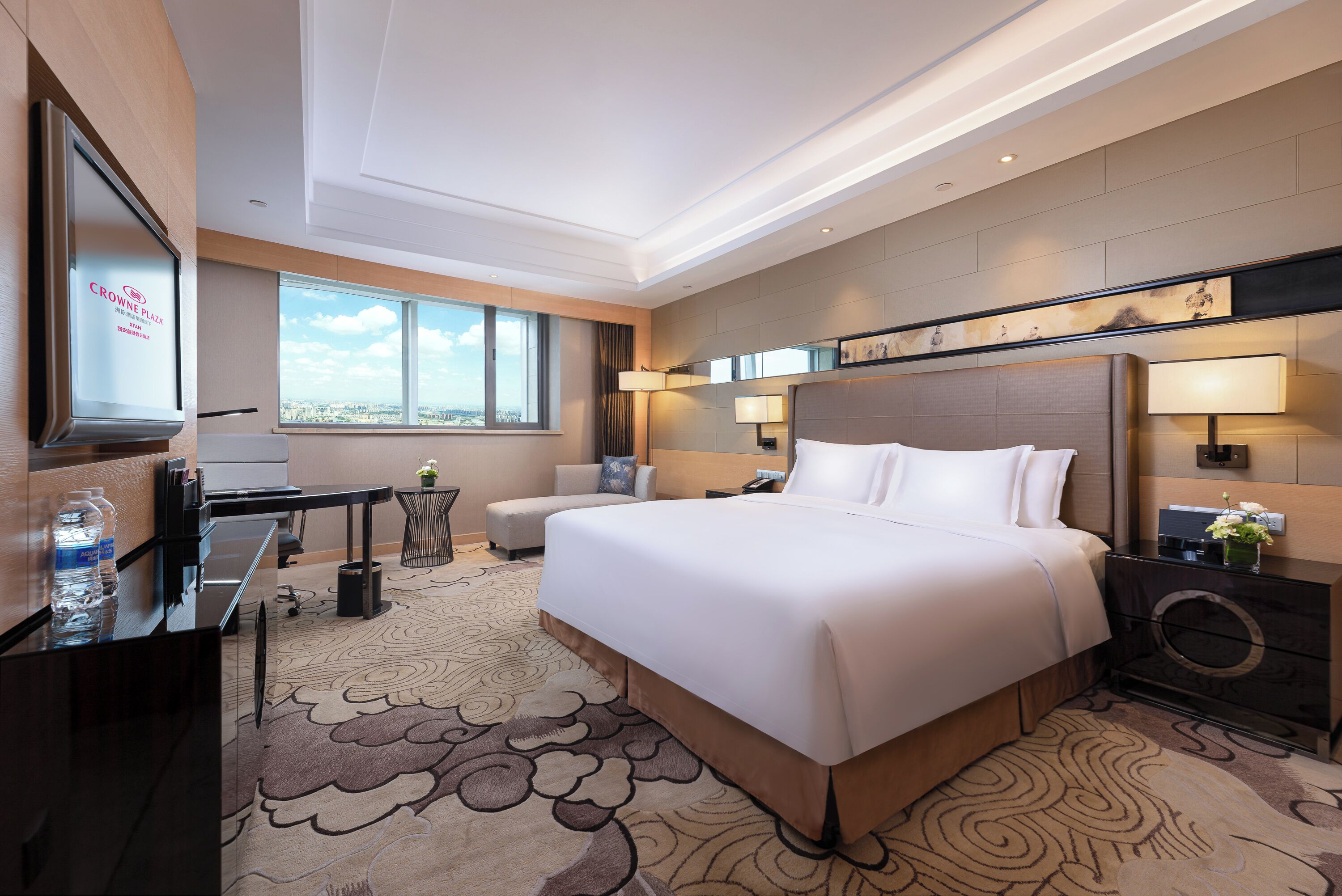 A hotel room with a large bed, a desk, a chair, a TV, and a view of the city.