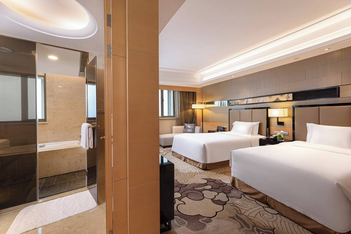 A hotel room with a glass shower enclosure, two beds, a bedside table with a lamp, and a large mirror.