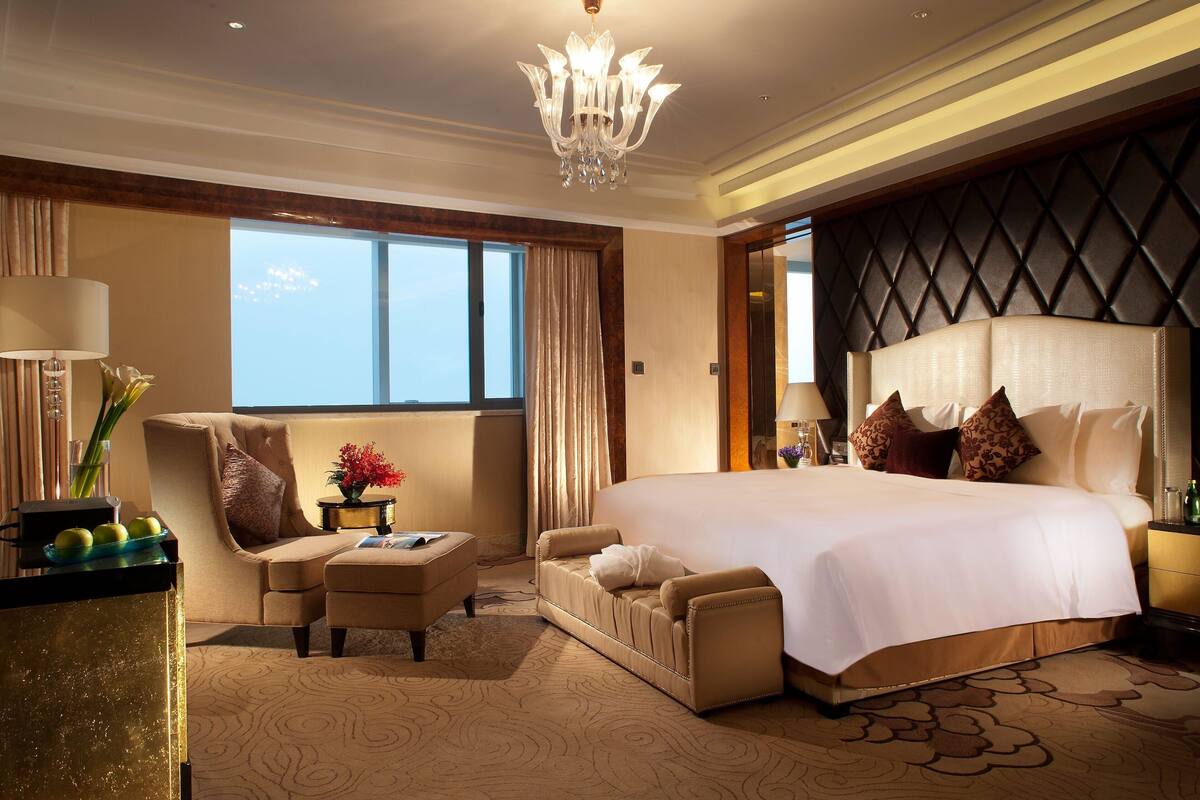 A spacious hotel room with a large bed, a sitting area with a sofa and armchair, a desk with a lamp, and a view of the sky through large windows.