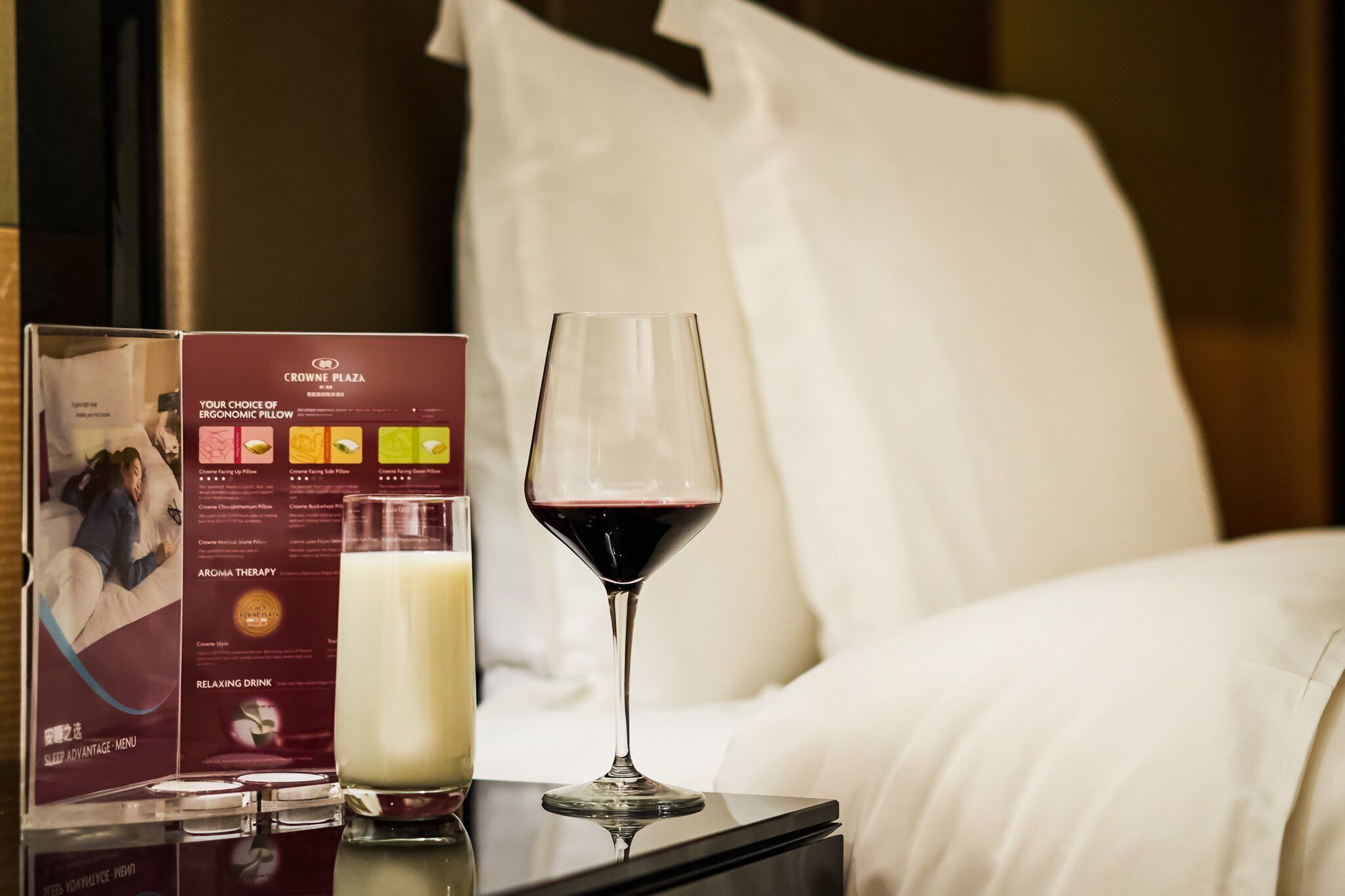 Una habitación de hotel con una cama, una copa de vino tinto, una botella de leche y un folleto sobre una mesita de noche.